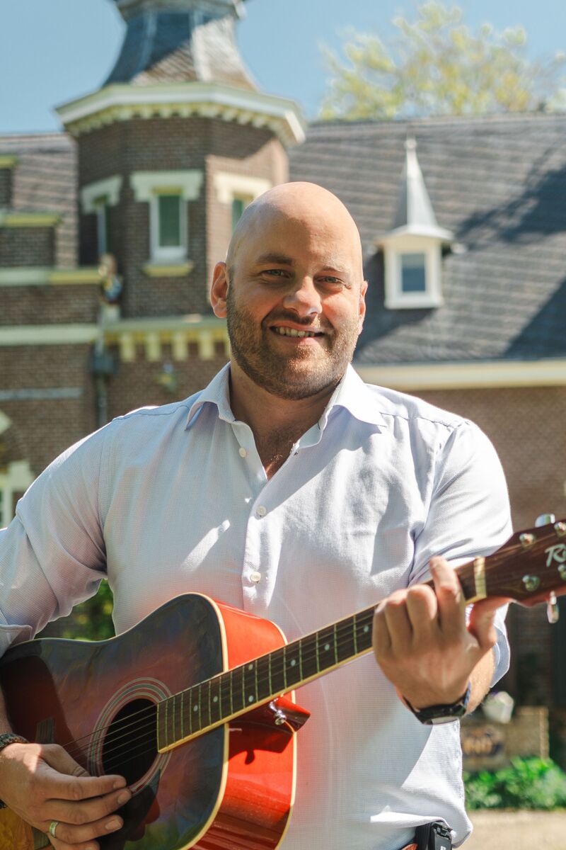 Portret foto van raadslid Bauke Beert Keizer met een rode gitaar voor een oud gebouw.