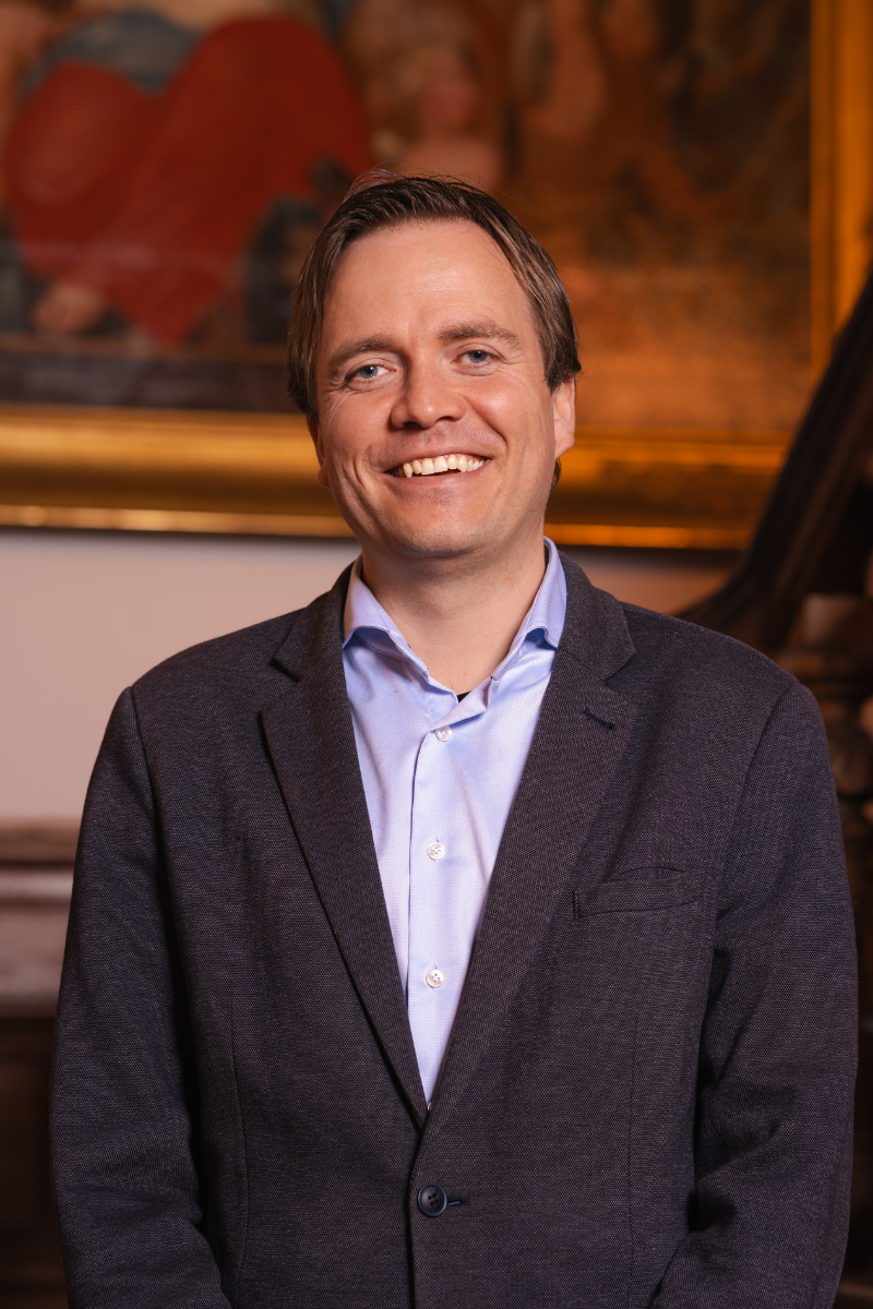 Foto van Johan Magré voor een houten trap en een schilderij. Statig portret.