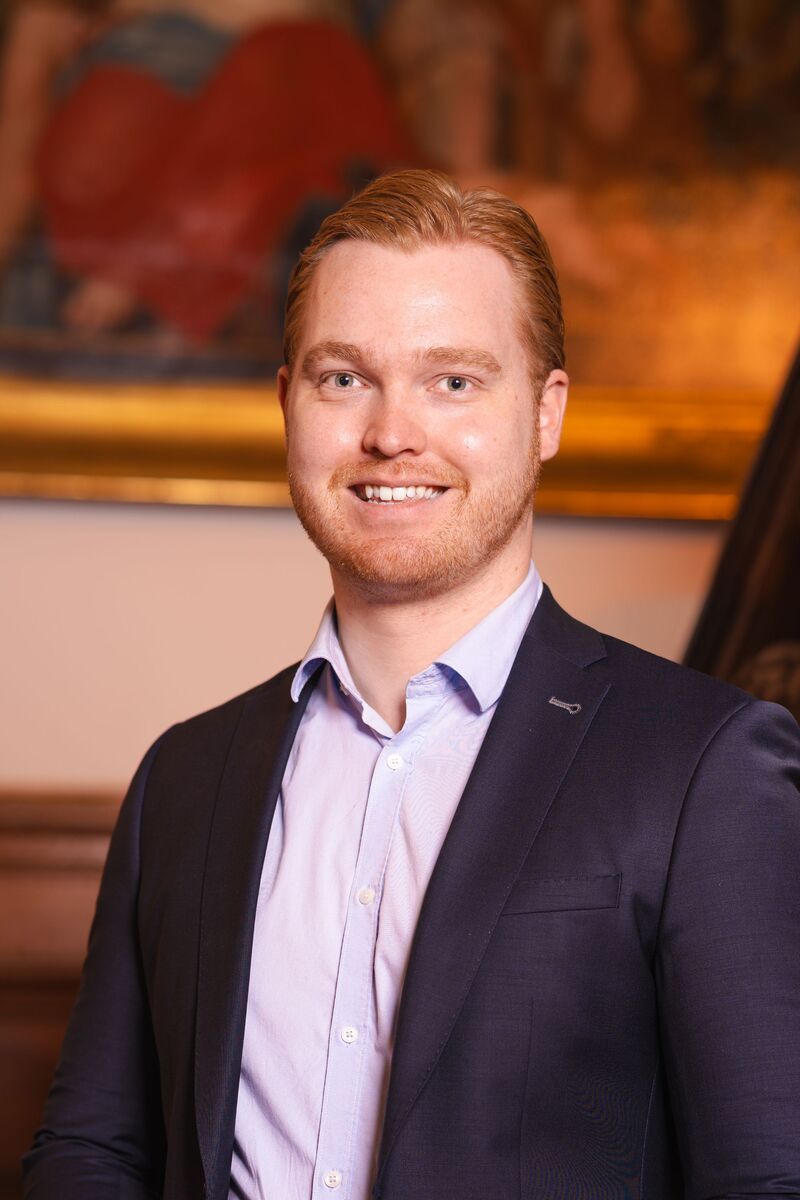 portretfoto van raadslid Justin voor een houten trap met een schilderij er achter. Een statig portret.