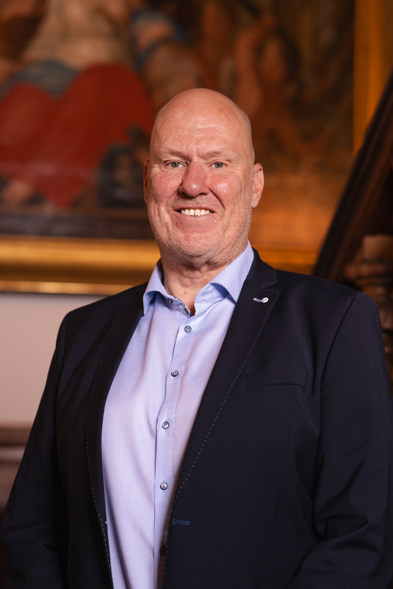 Portret foto van raadslid Dirk voor een houten trap met een schilderij op de achtergrond. Een statig portret.