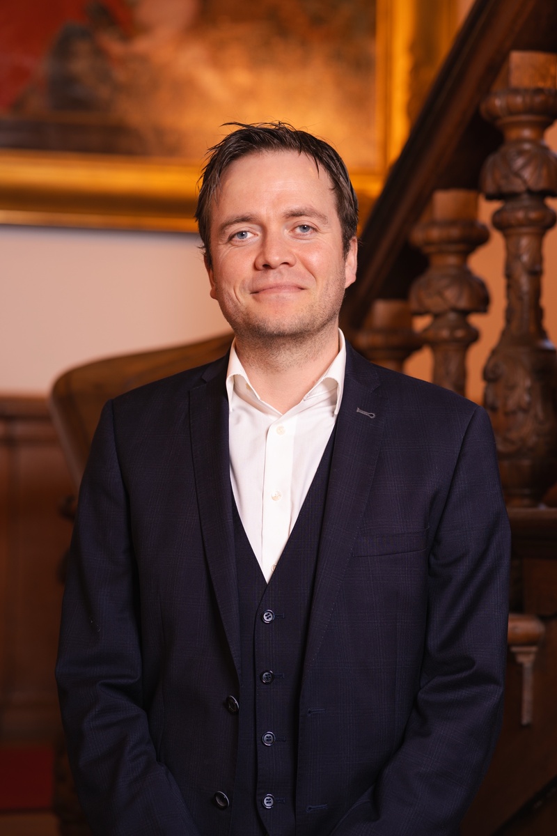Foto van Johan Magré voor een houten trap en een schilderij. Statig portret.