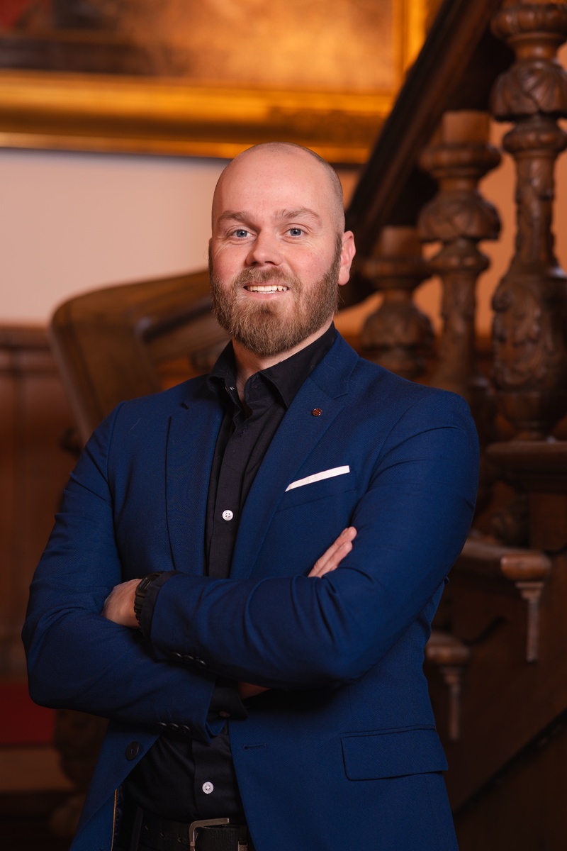 Portret foto van raadslid Kevin voor een houten trap met een schilderij op de achtergrond. Een statig portret.