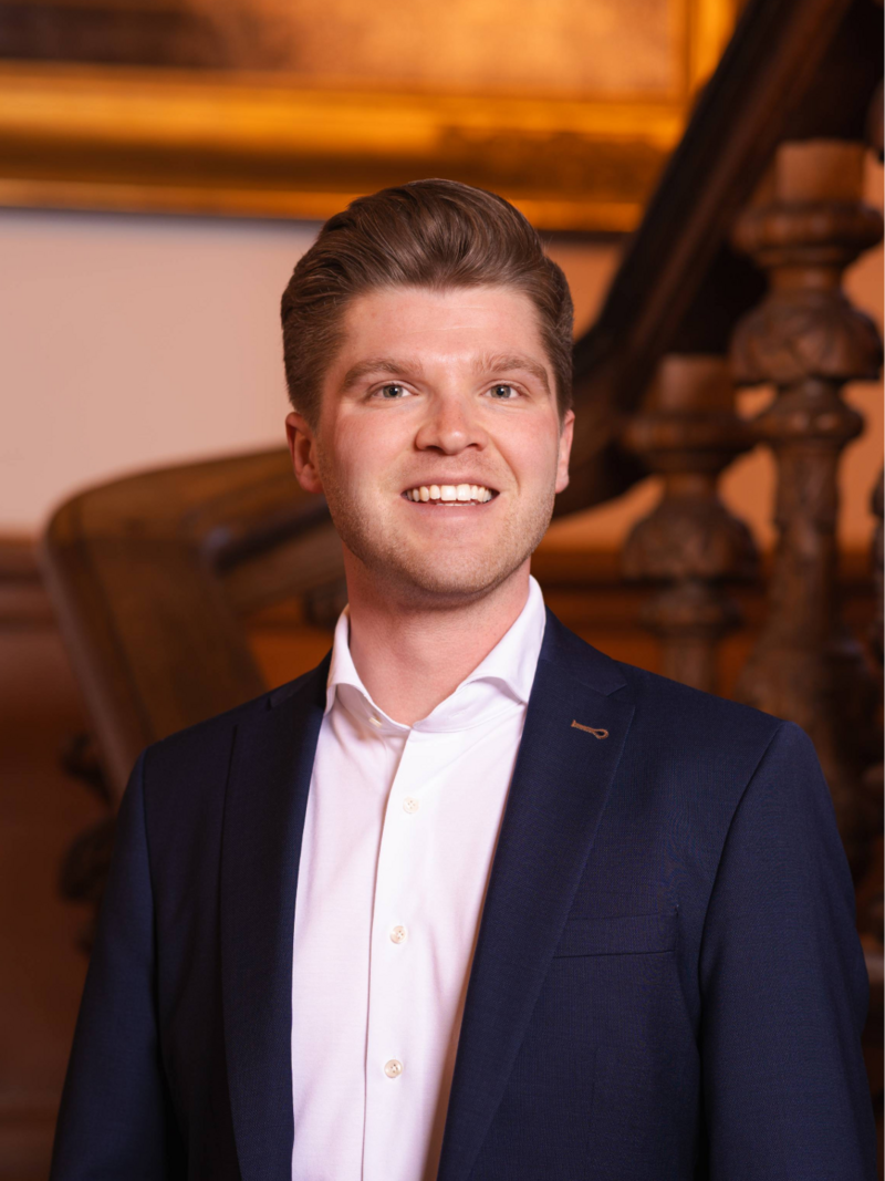Portret foto van raadslid Lennart voor een houten trap met een schilderij op de achtergrond. Een statig portret.