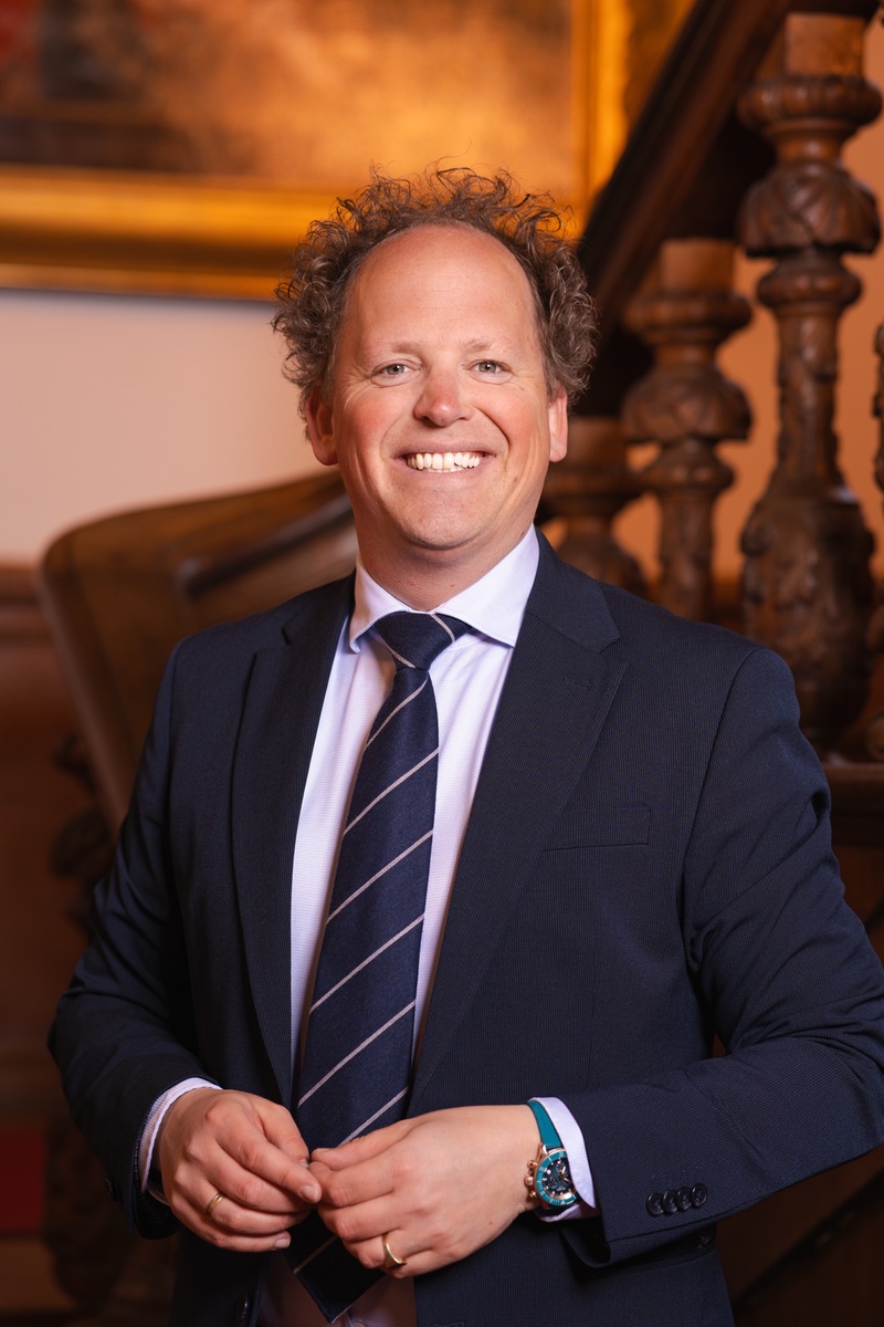 Portret foto van raadslid Abel voor een houten trap met een schilderij op de achtergrond. Een statig portret.