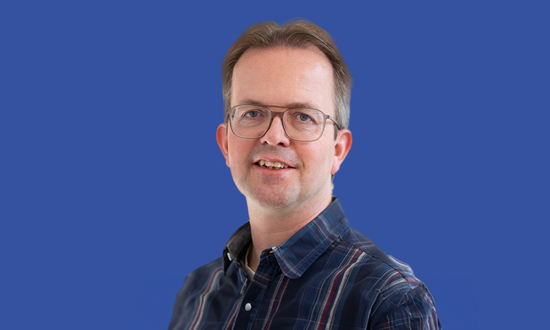 Foto van Christiaan Okkema in een gestreepte, donkerblauwe overhemd met rode lijnen. Voor een blauwe achtergrond.
