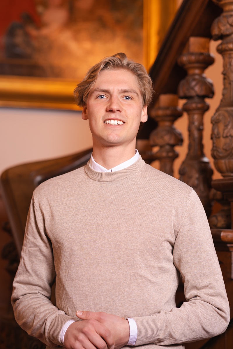 Portret foto van raadslid Thomas voor een houten trap met een schilderij op de achtergrond. Een statig portret.