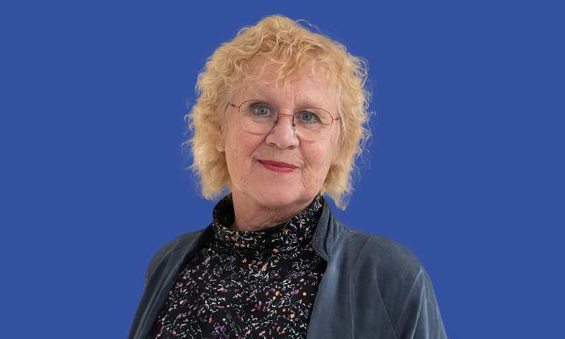 Foto van Riemy Spithoff, met een blauwe colbert en een gestipte zwarte blouse. Voor een blauwe achtergrond