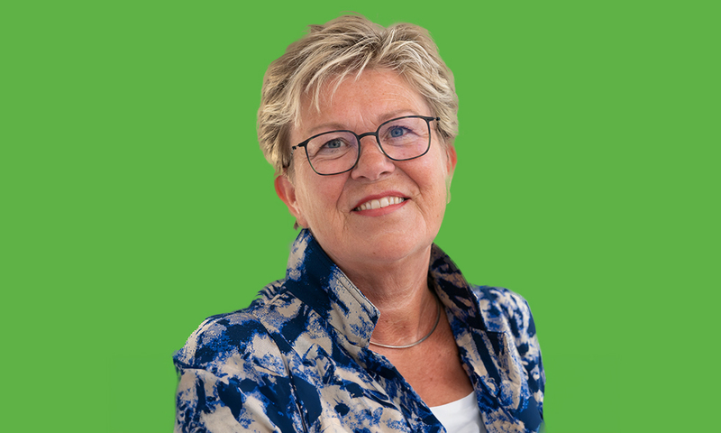 Een foto van Jannie Atsma, met een blauw-wit jasje aan en een wit shirt. Voor een groene achtergrond
