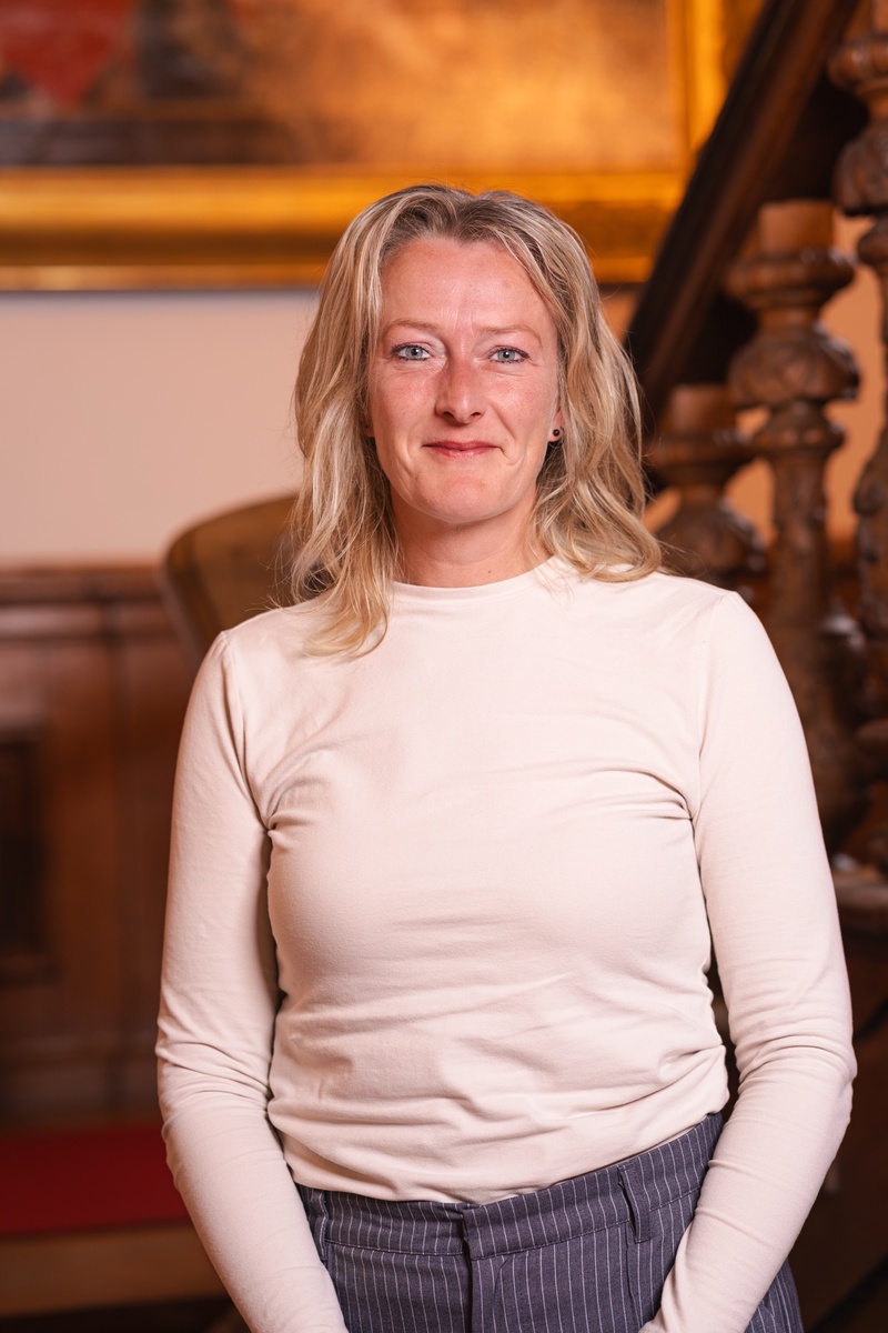 Portret foto van raadslid Janneke voor een houten trap met een schilderij op de achtergrond. Een statig portret.