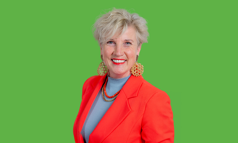 Een foto van Fokelien van der Meulen met een fel oranje blazer en blauwe trui. Voor een groene achtergrond.