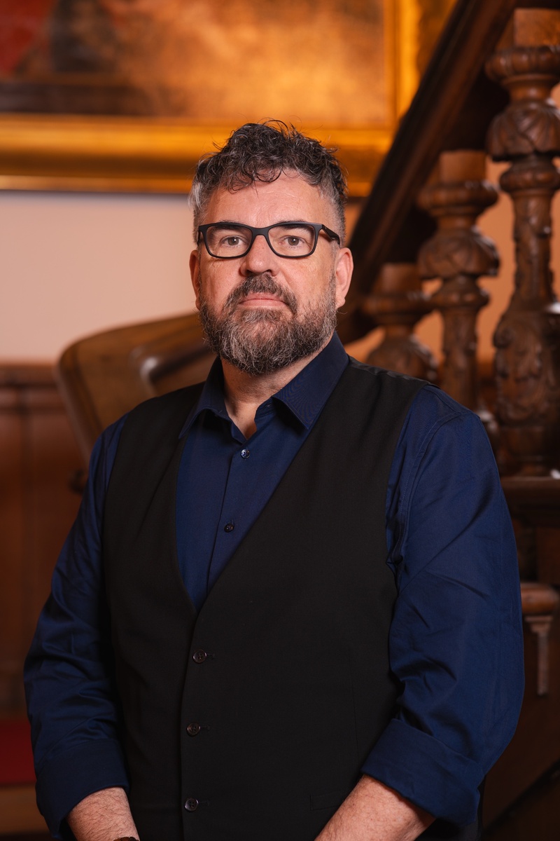 Portret foto van raadslid Johannes voor een houten trap met een schilderij op de achtergrond. Een statig portret.