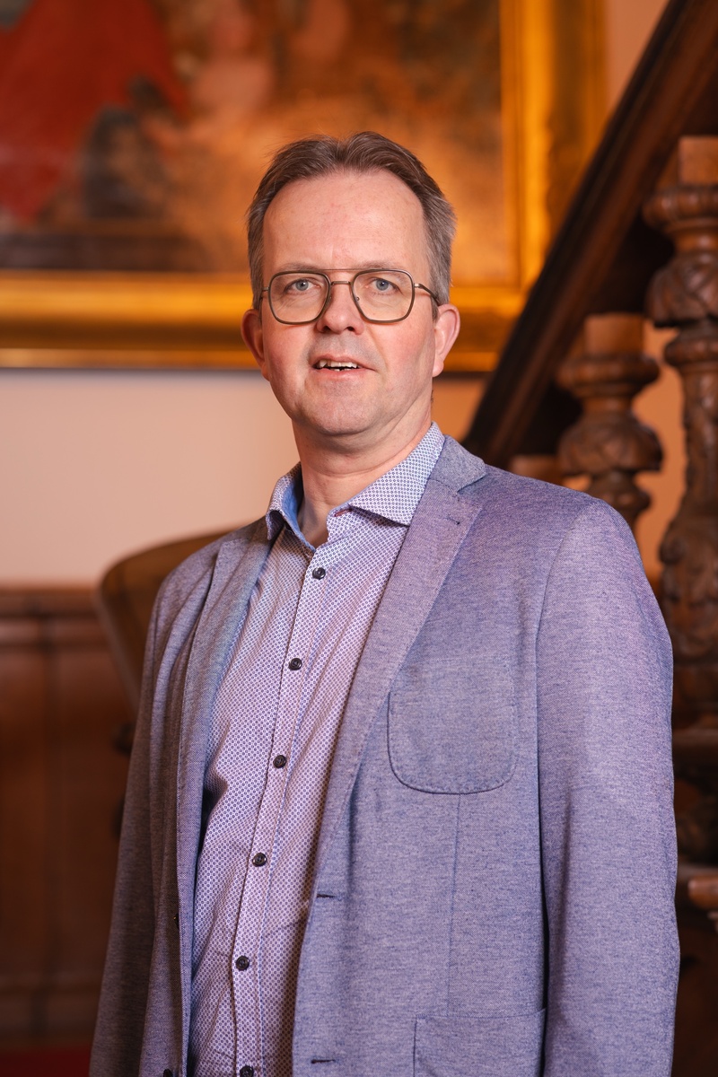 Portret foto van raadslid Christiaan voor een houten trap met een schilderij op de achtergrond. Een statig portret.