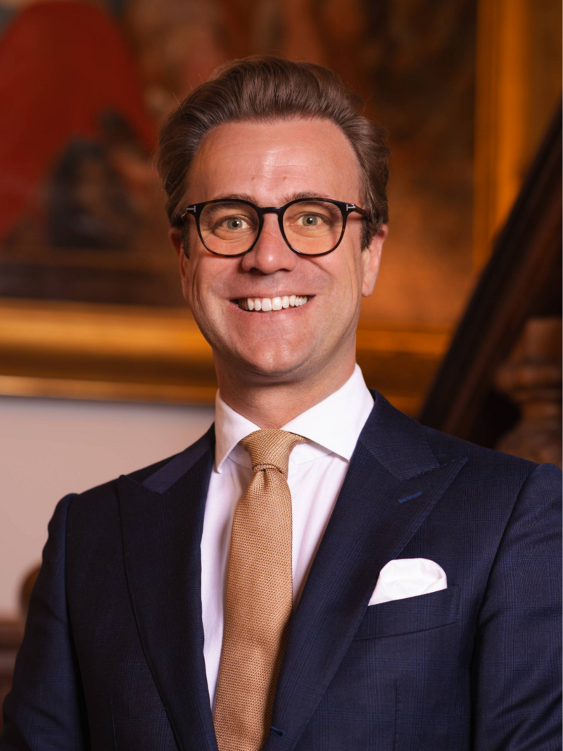 Portret foto van raadslid Ruthger voor een houten trap met een schilderij op de achtergrond. Een statig portret.